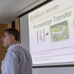 A man with a slide titled Knowledge Transfer is projected on a screen behing him.